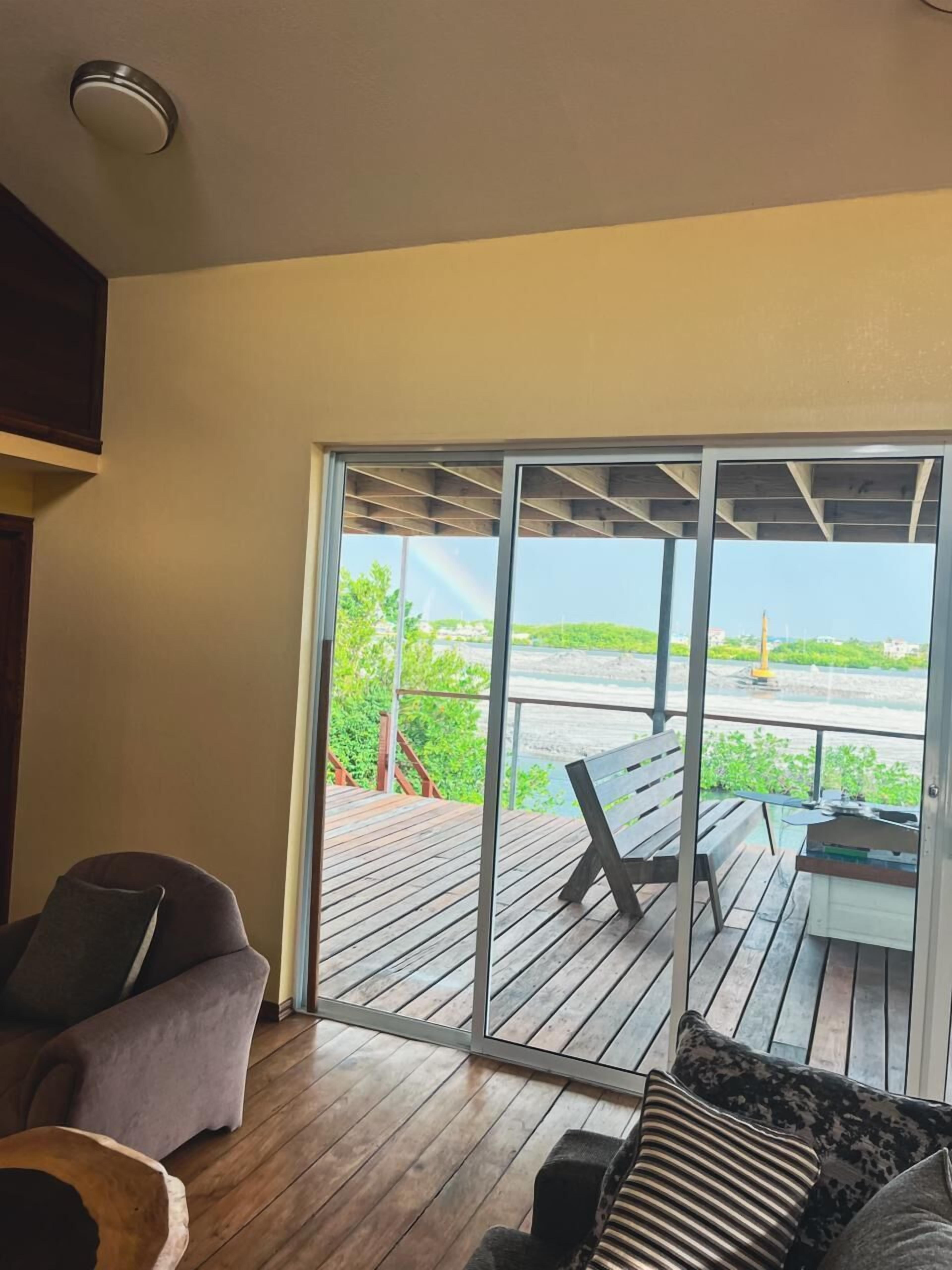 A room with a wooden deck, a sliding glass door leading to an outdoor area with a table and chairs, and a cushioned seating area.