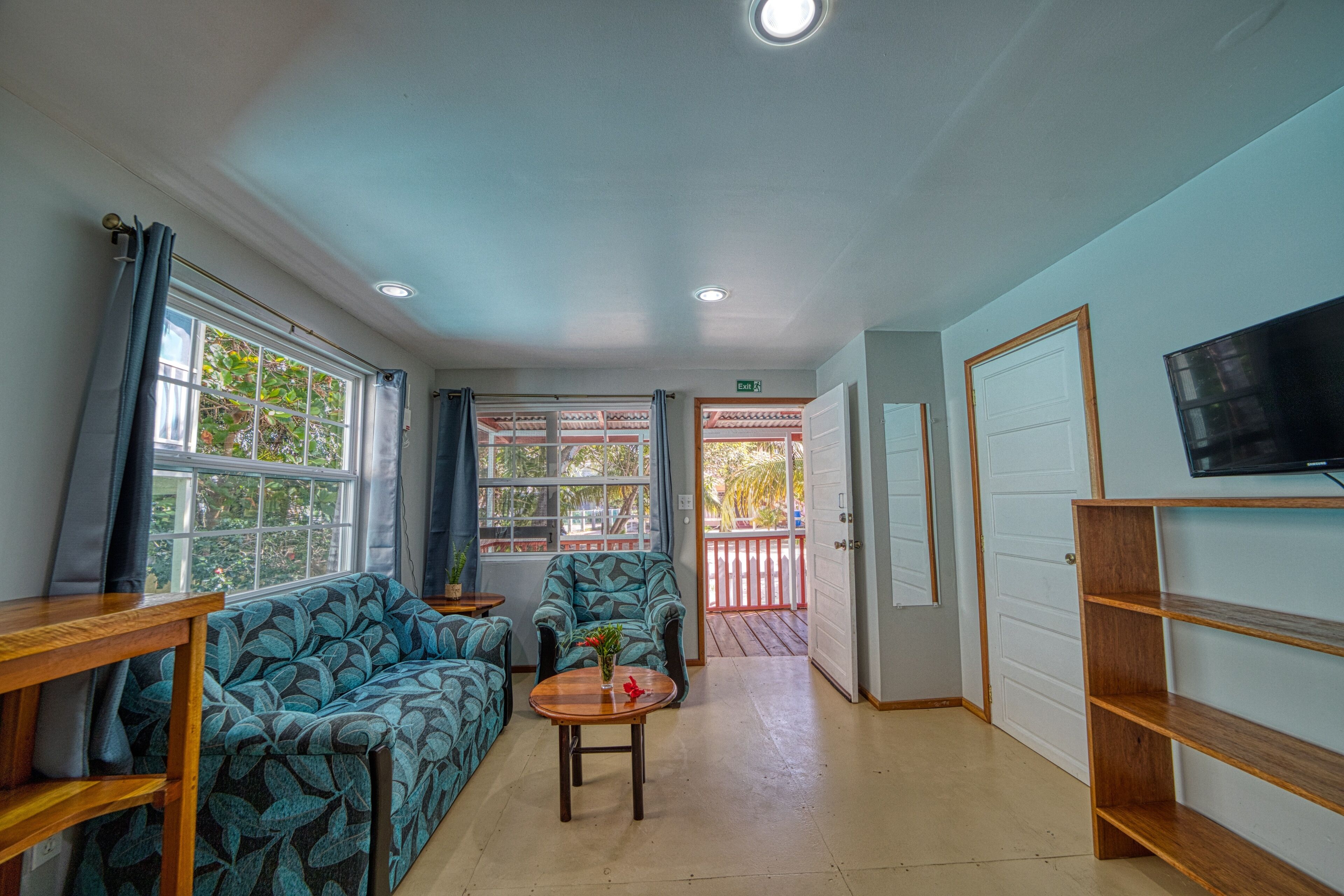 A room with a sofa, armchair, and a small table with a vase. There is a TV mounted on the wall and a door leading outside.