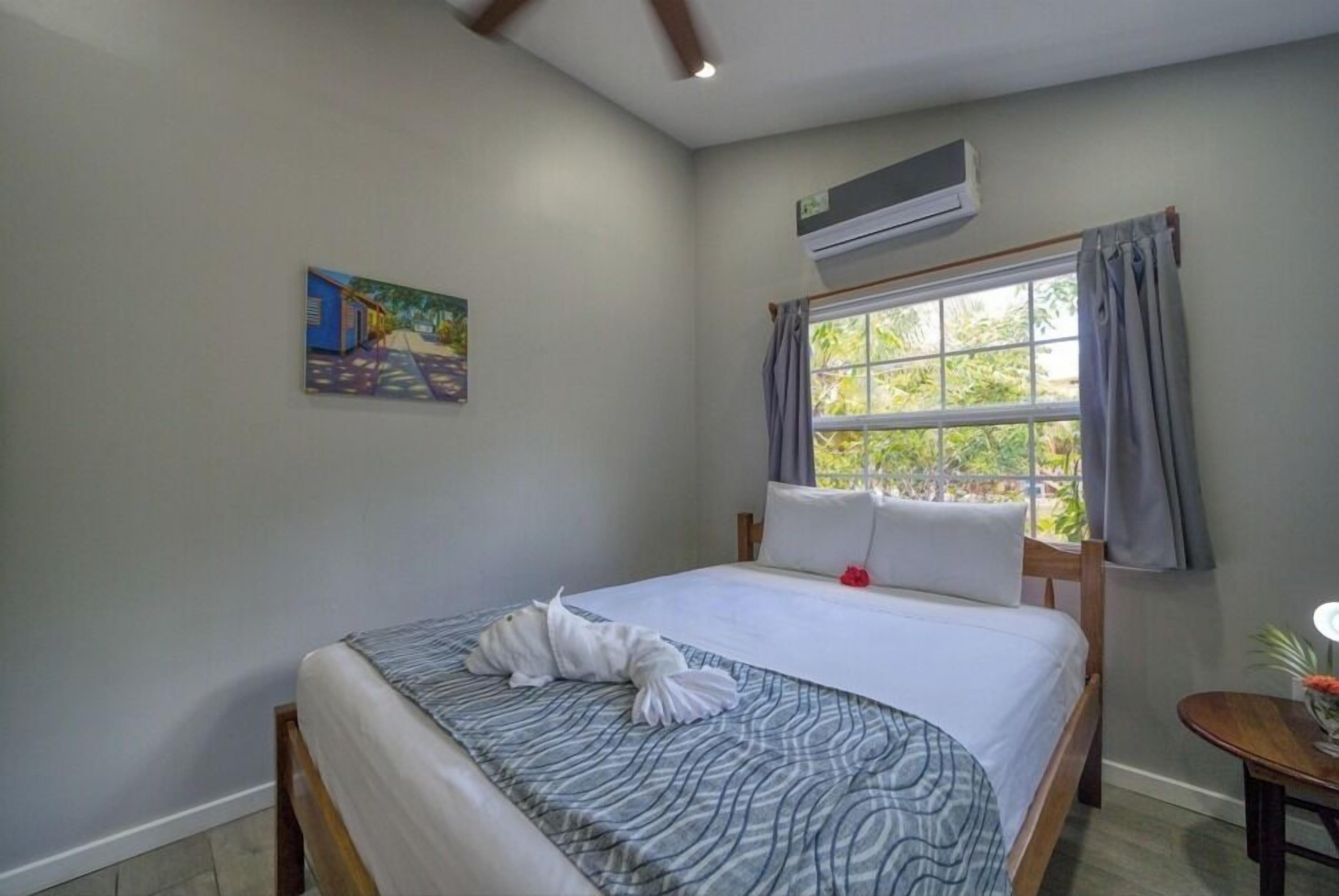 A neatly made bed with a patterned bedspread, a painting on the wall, an air conditioning unit, and a window with curtains.