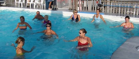 Une piscine où l’on voit des personnes dans l’eau, entourée d’une terrasse en bois et de chaises en plastique blanches.