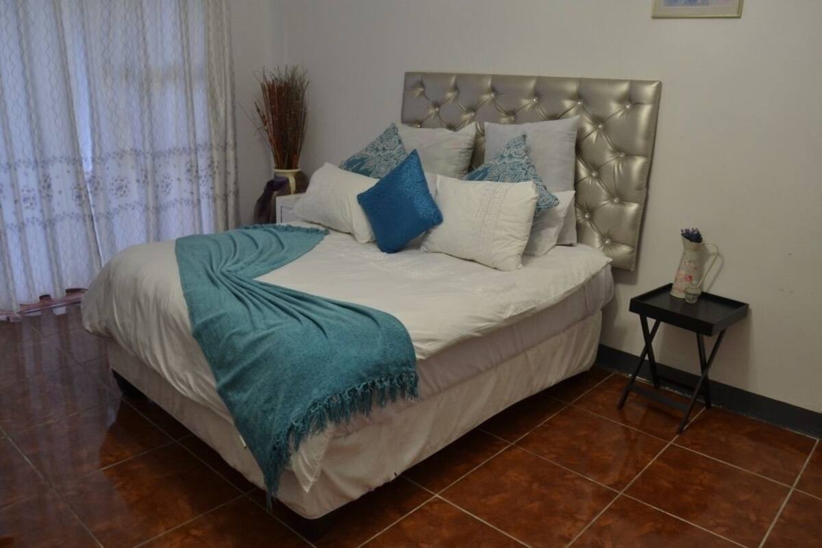 Een slaapkamer met een geborduurde hoofdbord, een bed met wit en blauw beddengoed, een blauwgroene plaid, een klein nachtkastje met een vaas, en een raam met dunne gordijnen.