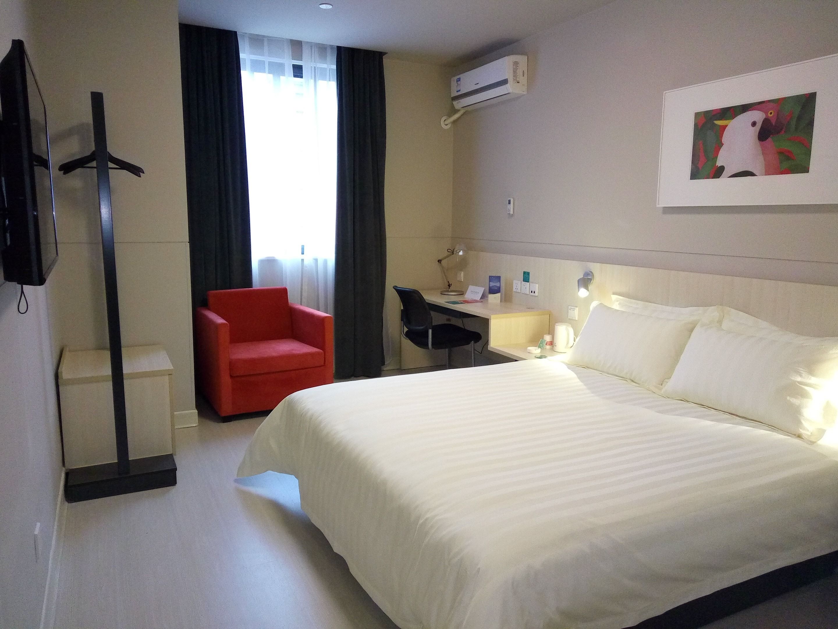 A hotel room with a bed, a desk, a chair, an air conditioner, and a painting on the wall.