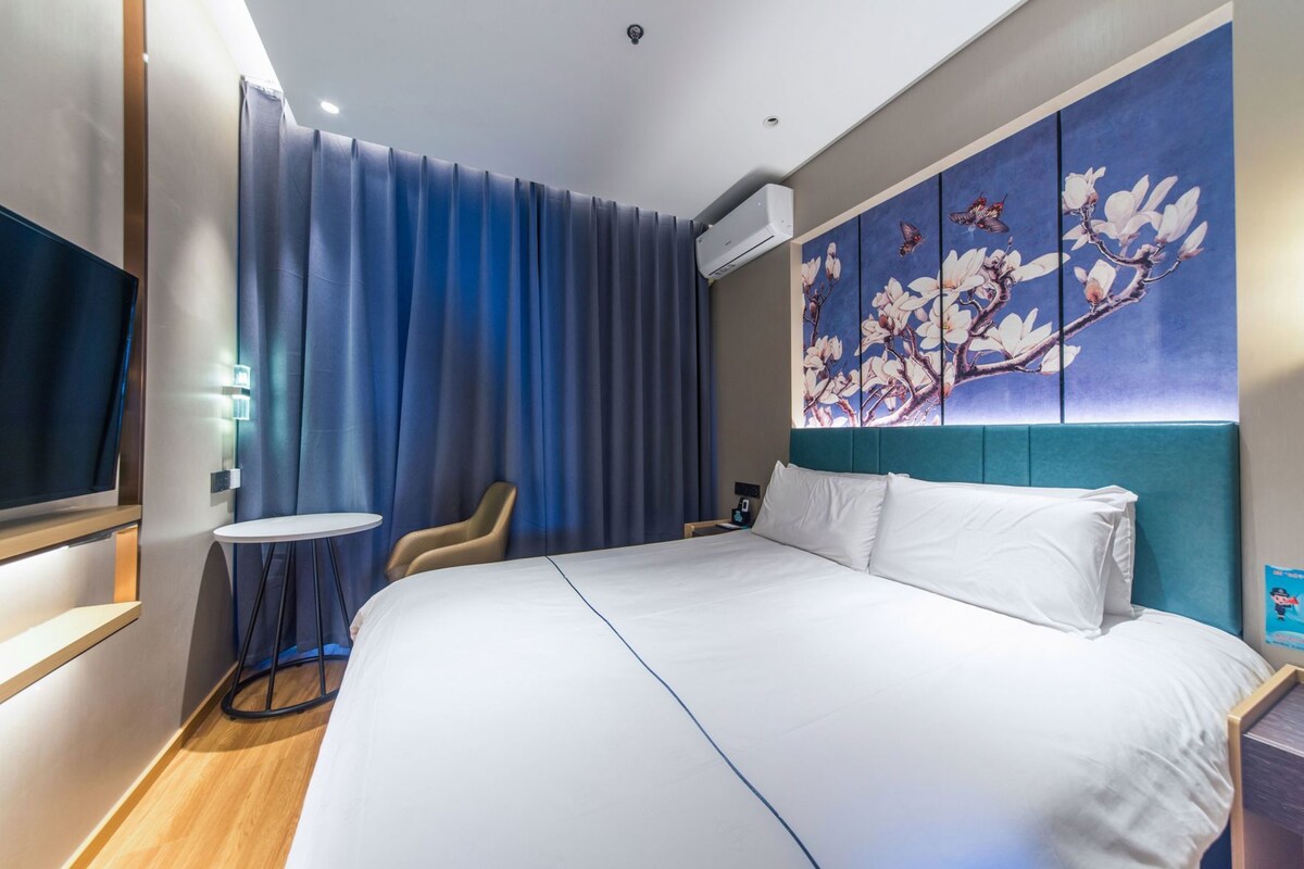 Een hotelkamer met een groot bed, een flatscreen televisie, een klein rond tafeltje, een stoel en een wandgemonteerde airconditioner.