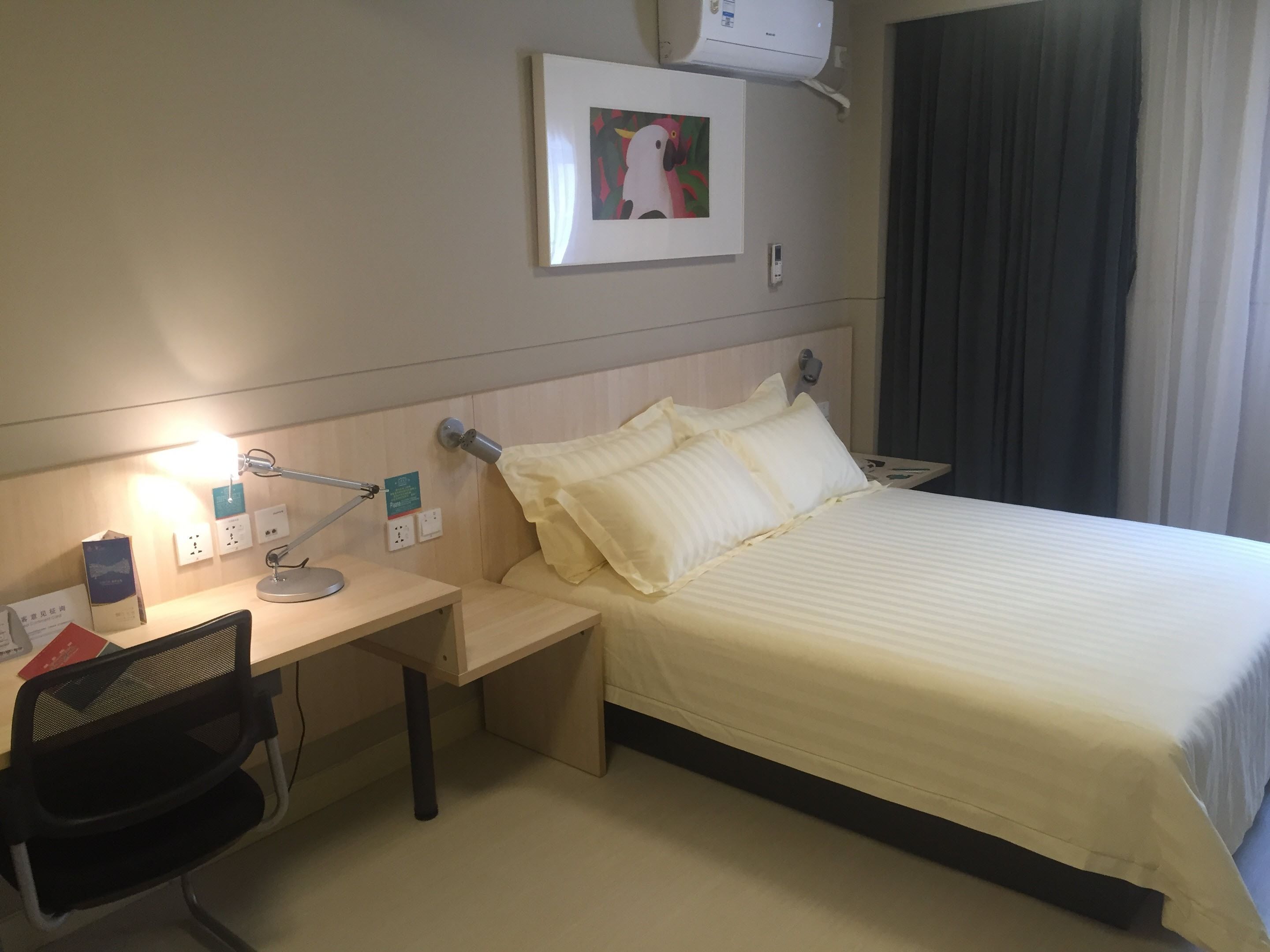 A hotel room with a bed, desk, chair, and a wall-mounted air conditioner.