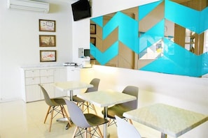A dining area with tables and chairs, a kitchen area with a refrigerator and microwave, and a wall with geometric patterns.