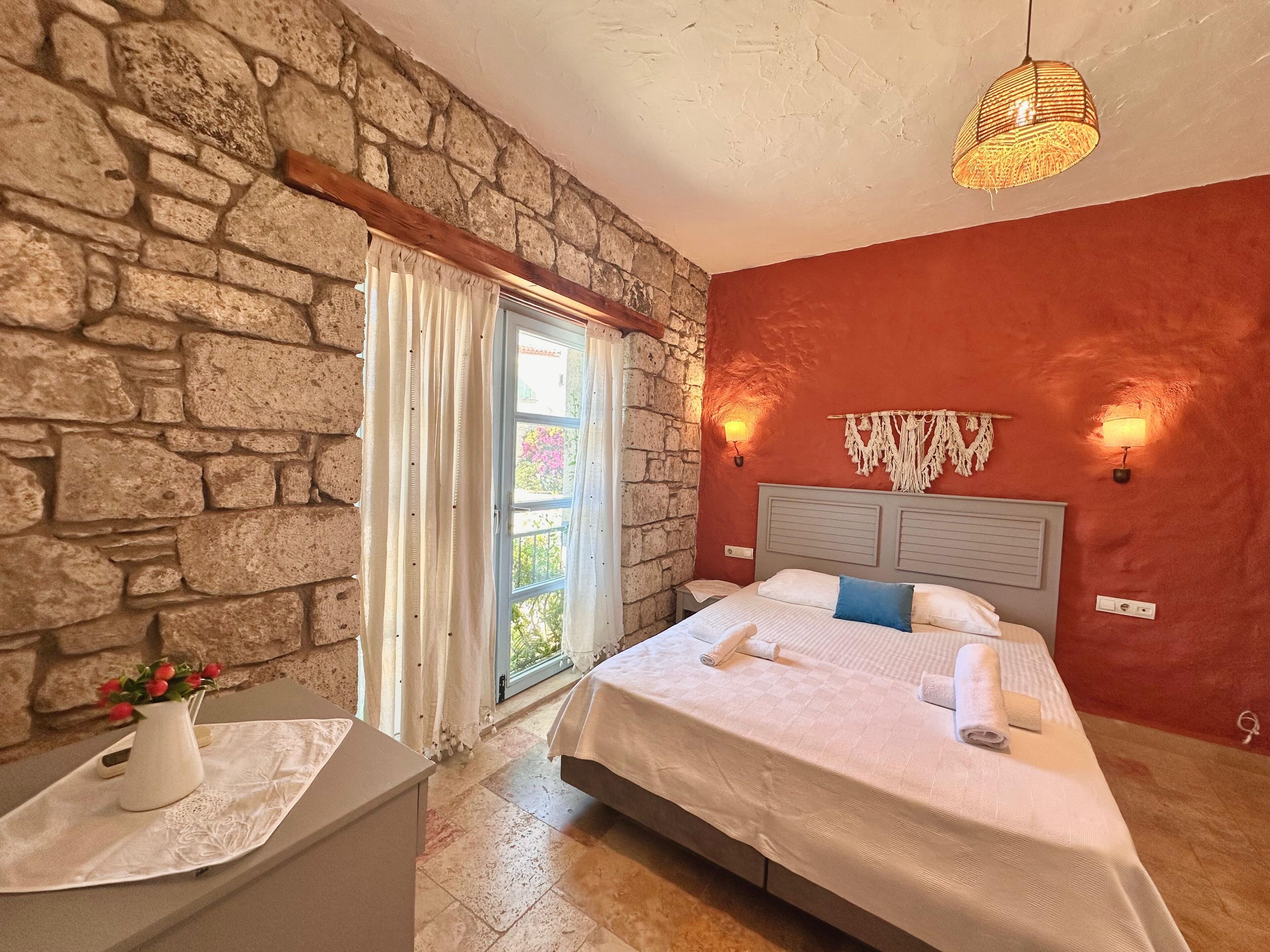 Ein Schlafzimmer mit Steinsandsteinwand, einem Bett mit weißer Bettwäsche, einem Nachttisch mit Blumenvase und einem Vorhangfenster.