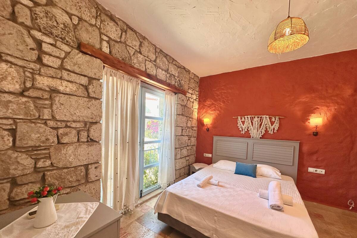 A bedroom with a stone wall, a bed with white bedding, a nightstand with a vase of flowers, and a window with curtains.