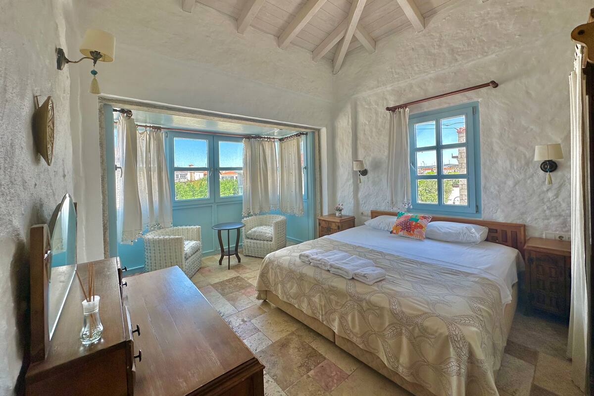 A bedroom with a bed, two bedside tables, a chair, a small table, and a window with curtains.