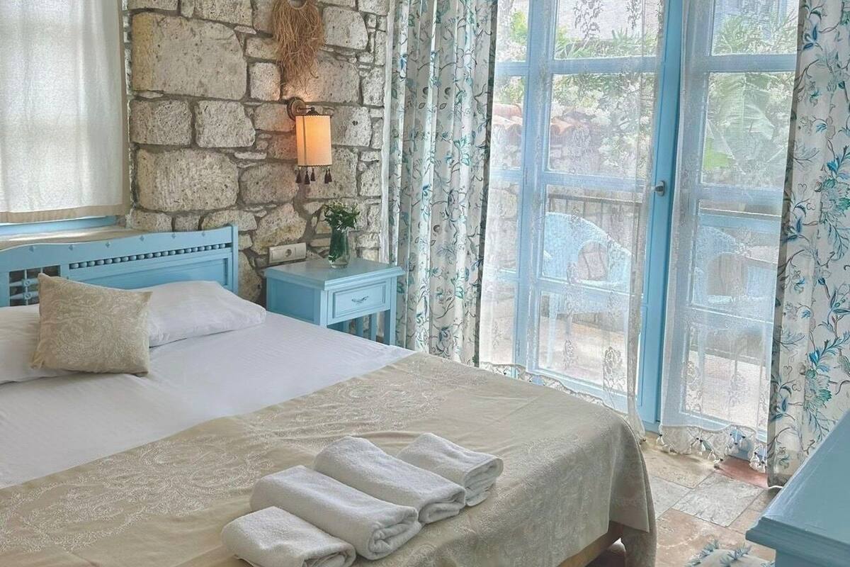 A bedroom with a stone wall, a bed with white linens, a nightstand with a lamp, and a view through a window with patterned curtains.