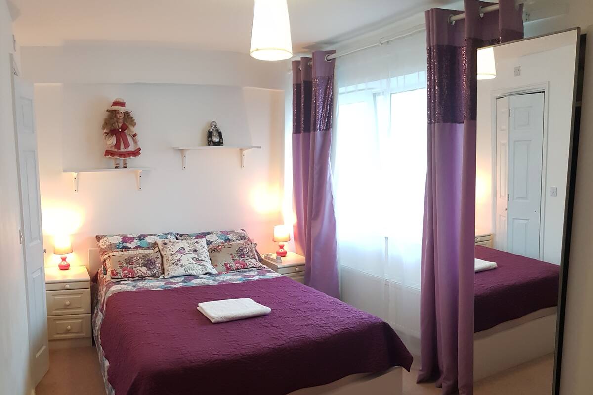 A bedroom with two beds, each with a purple bedspread and a white pillow. There are two bedside tables with lamps, a mirror, and a shelf with decorative items.