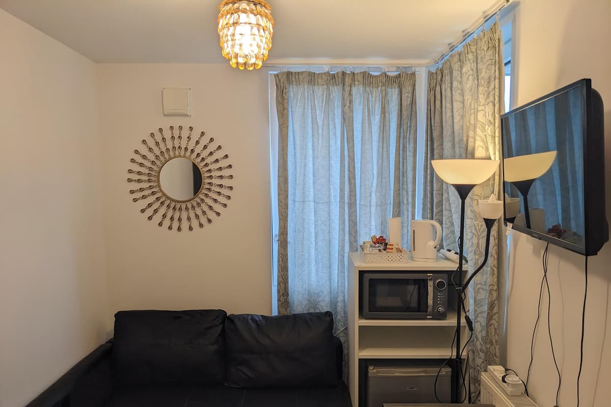 A compact living space with a black sofa, a small table, a microwave, a coffee maker, a lamp, and a mirror on the wall.
