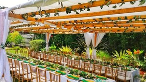Un espacio de comedor al aire libre con mesas dispuestas para una comida, rodeado de follaje y una estructura de pérgola.