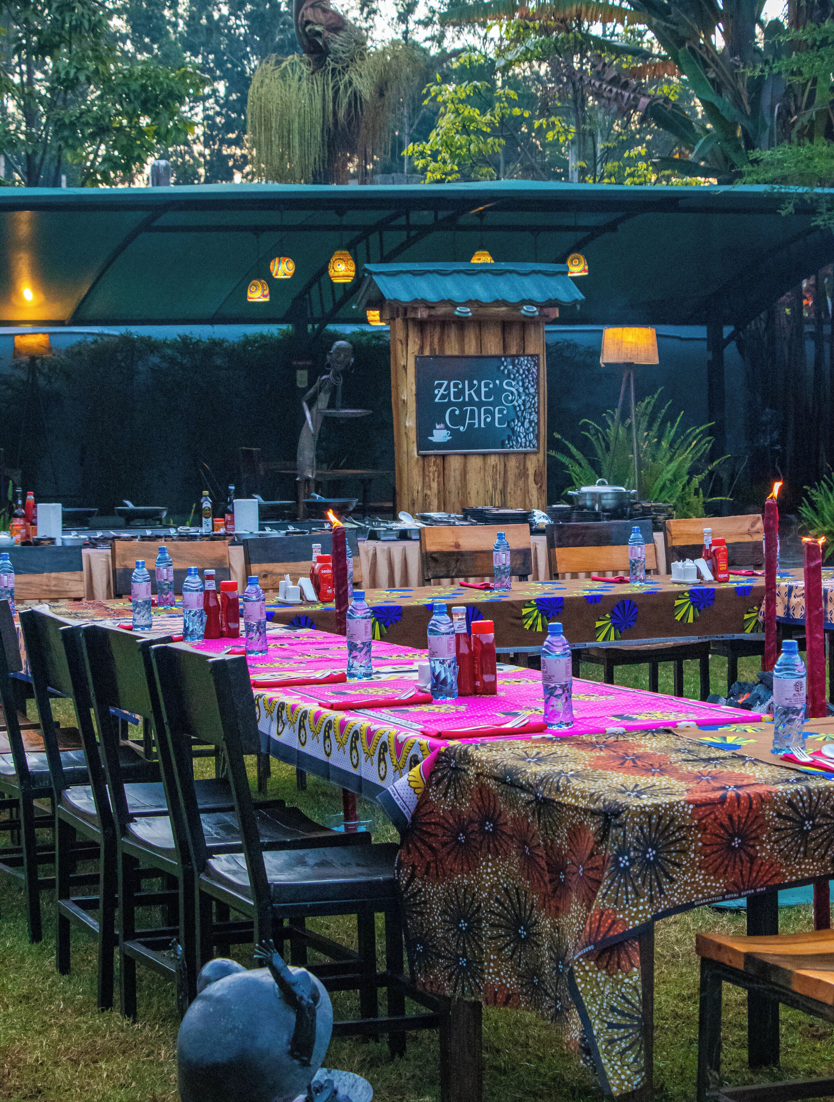 An outdoor dining area with tables set for a meal, a chef preparing food, and a sign that reads "Zeke's Cafe".