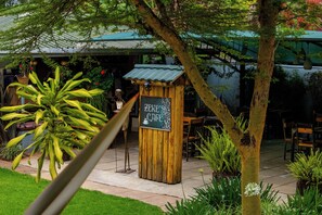 An outdoor seating area with a wooden sign reading "ZERE'S CAFE" and a variety of plants and trees. - Acacia Tree Lodge (Nairobi)