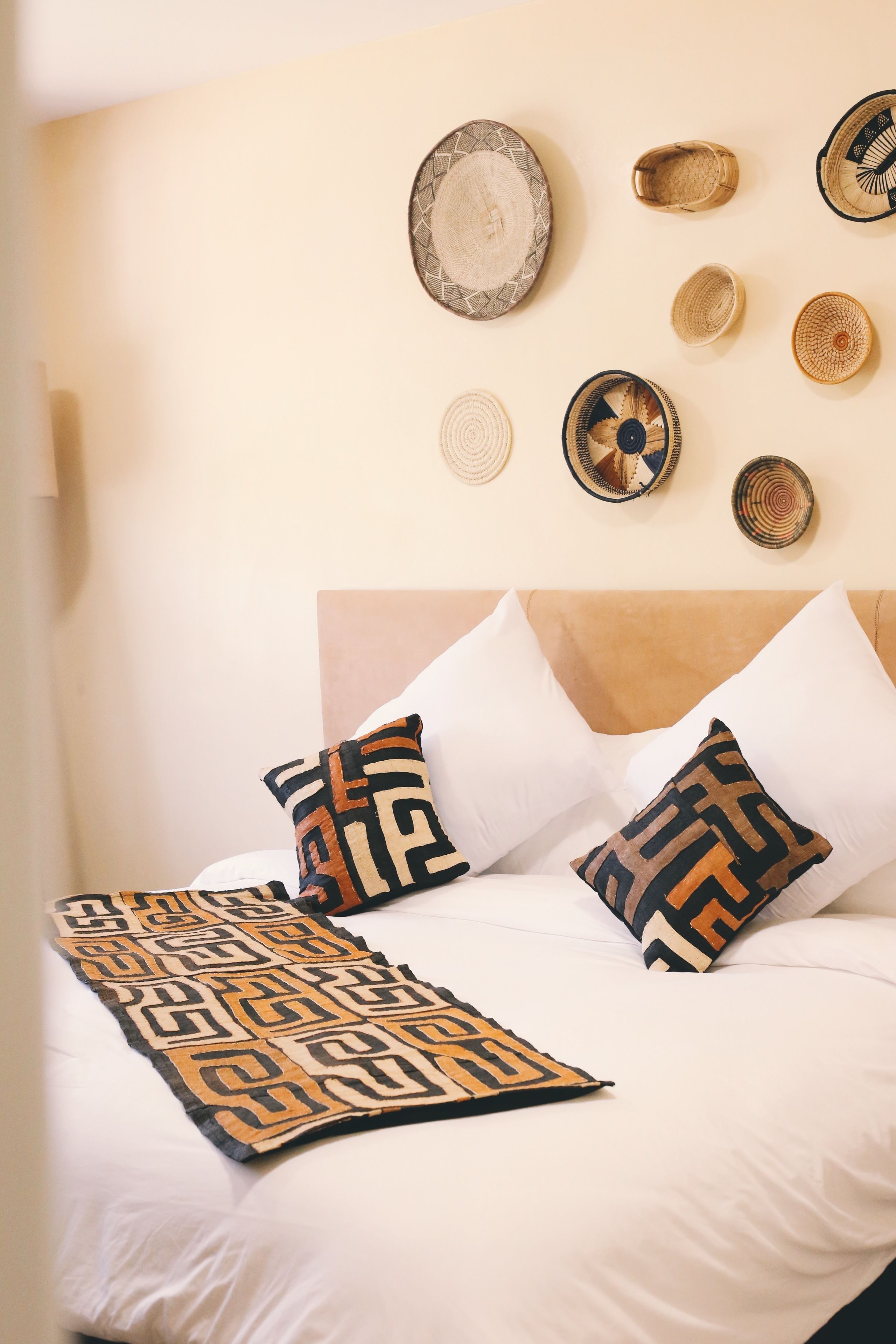 A bed with white bedding and patterned pillows, a headboard, and a wall decorated with woven baskets.