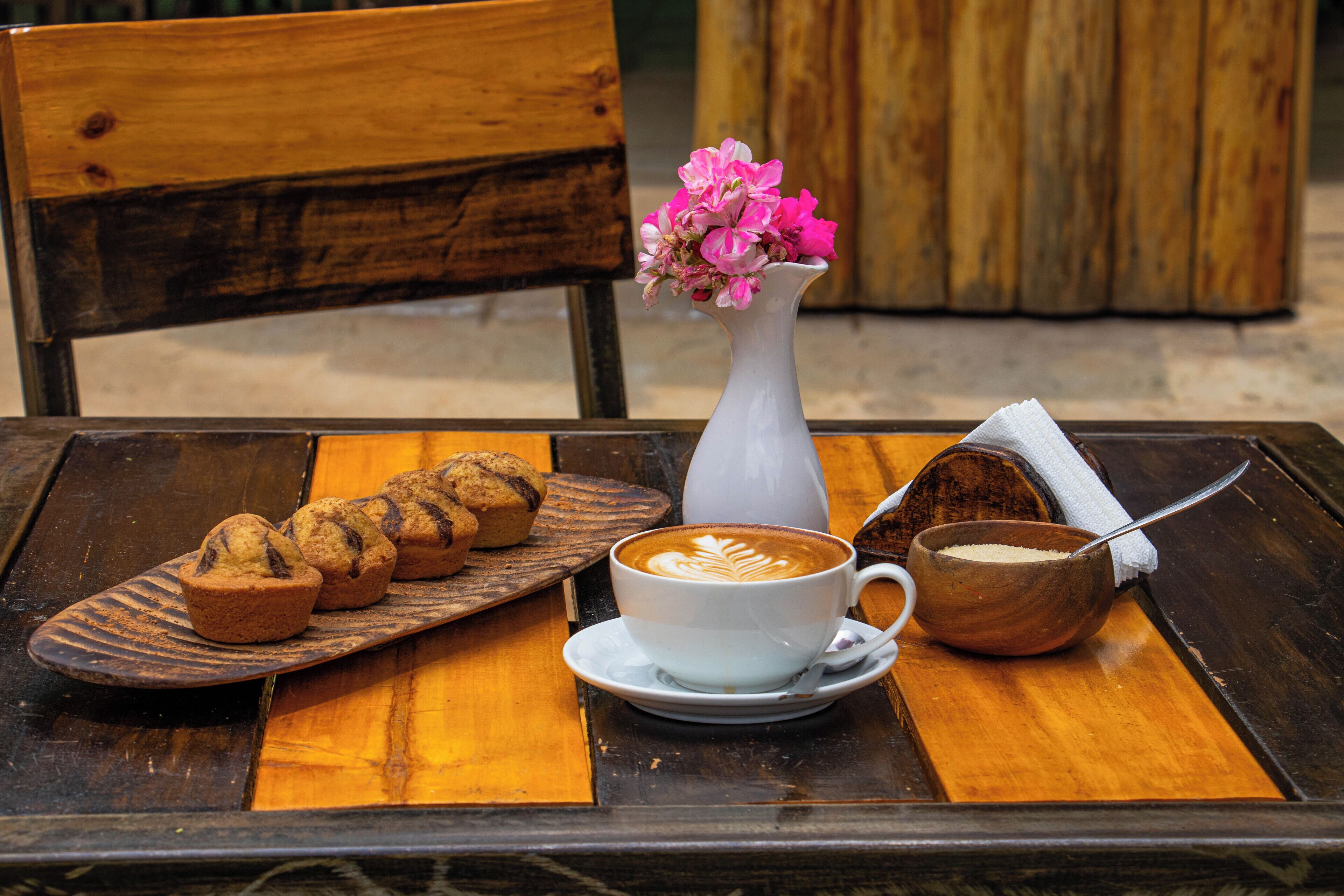 Ein Holztisch mit einer weißen Kaffeetasse, einer Holzschale, einem Teller mit Muffins und einer weißen Vase mit Blumen.
