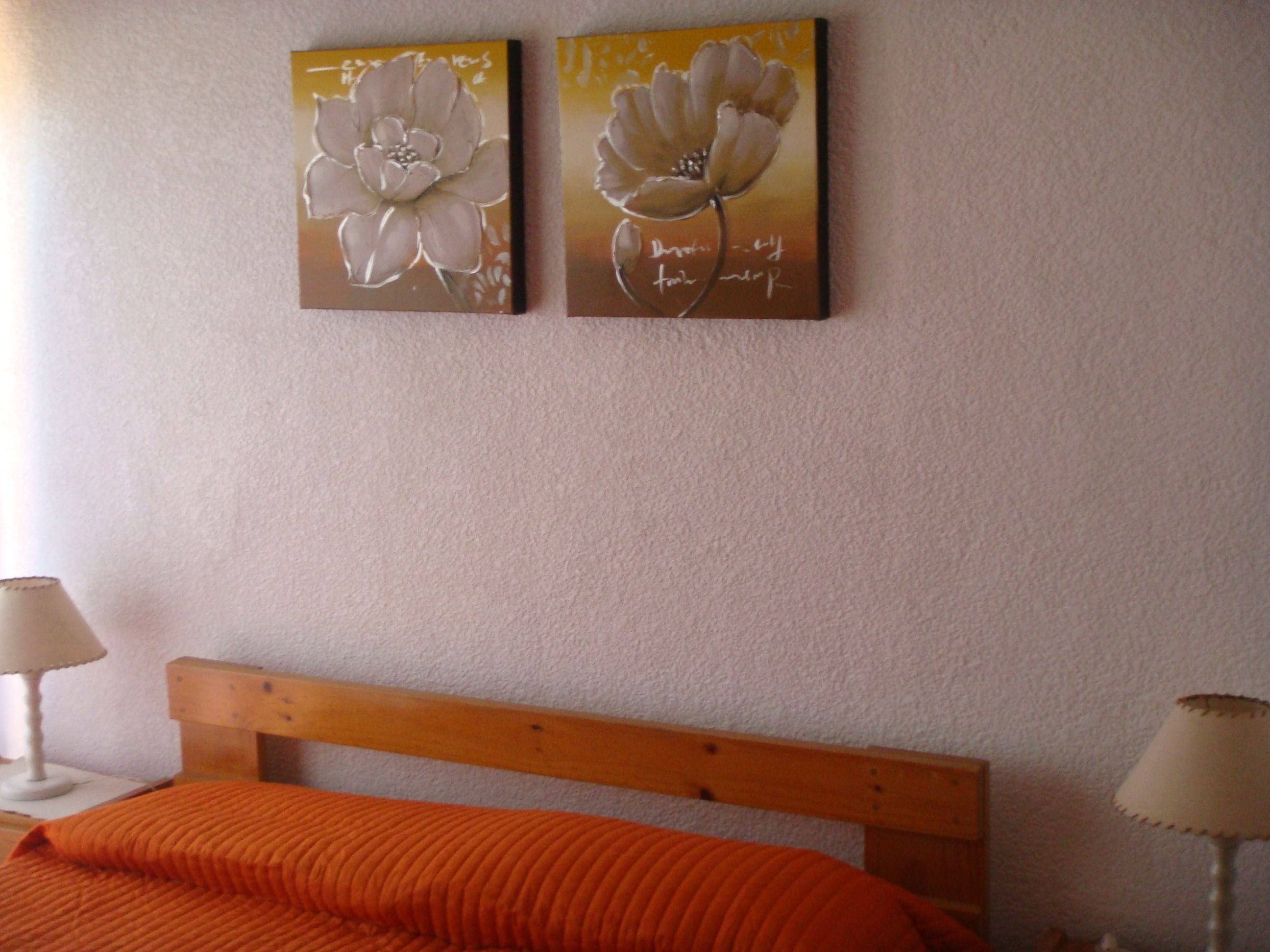 Ein Schlafzimmer mit einem Holz-Kopfteil, einer orangefarbenen Bettdecke und zwei an der Wand befestigten floralen Bildern.