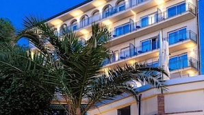 Um hotel com vários andares, fachada de pedra, uma palmeira e uma escadaria que leva à entrada.
