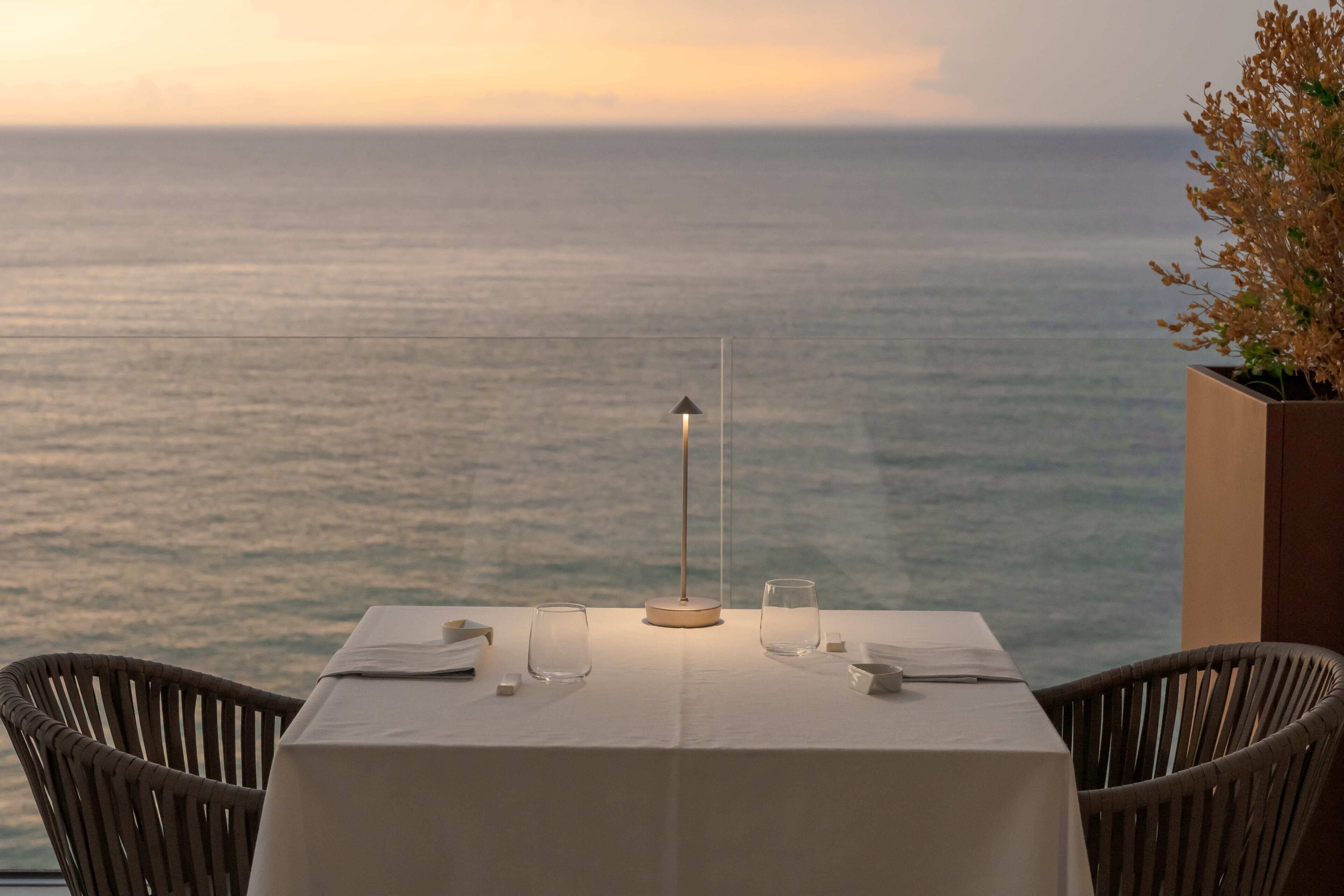 A table set for two with a view of the sea, a lamp, and a potted plant.