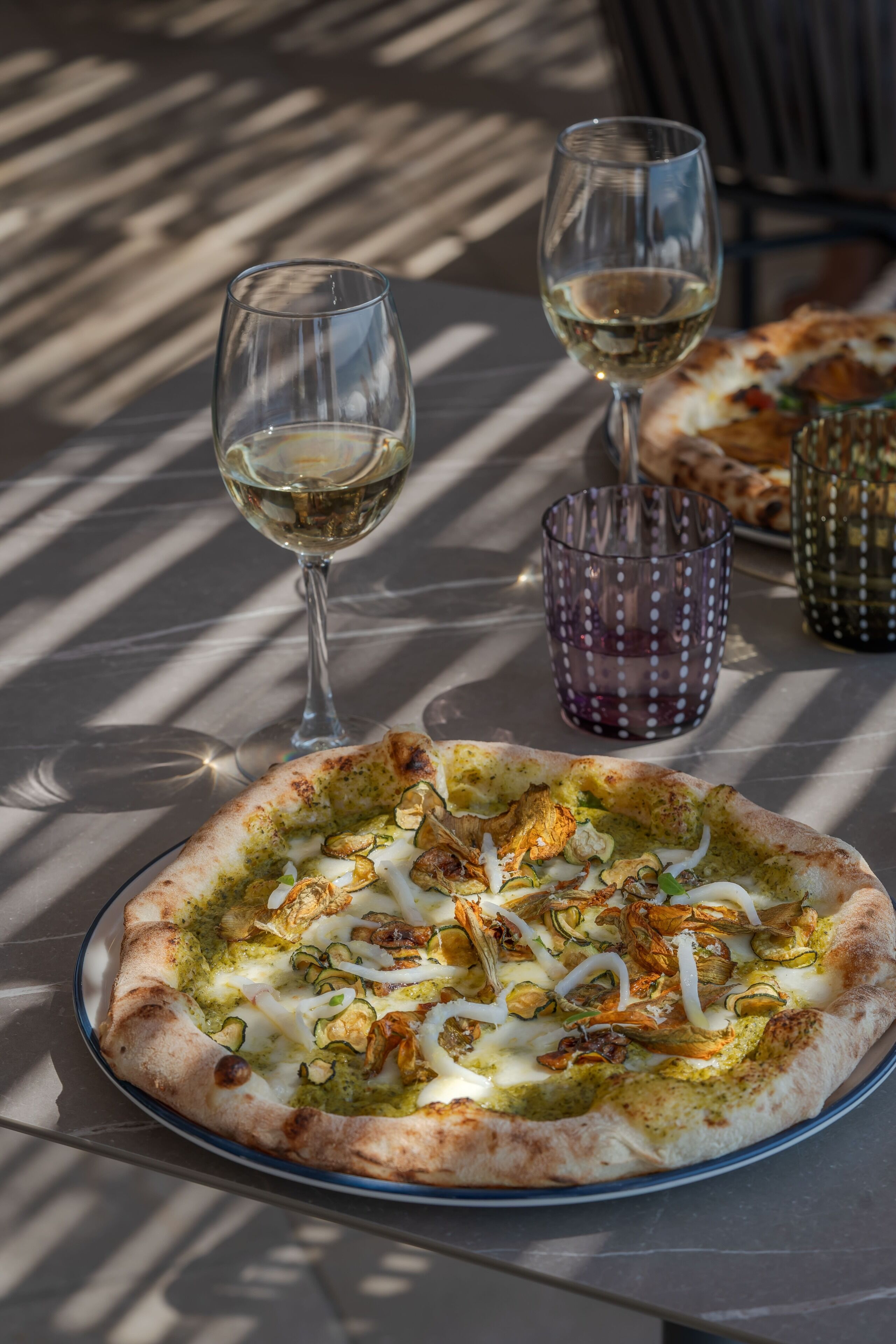 A pizza on a glass plate with a glass of white wine and a glass with a purple pattern.