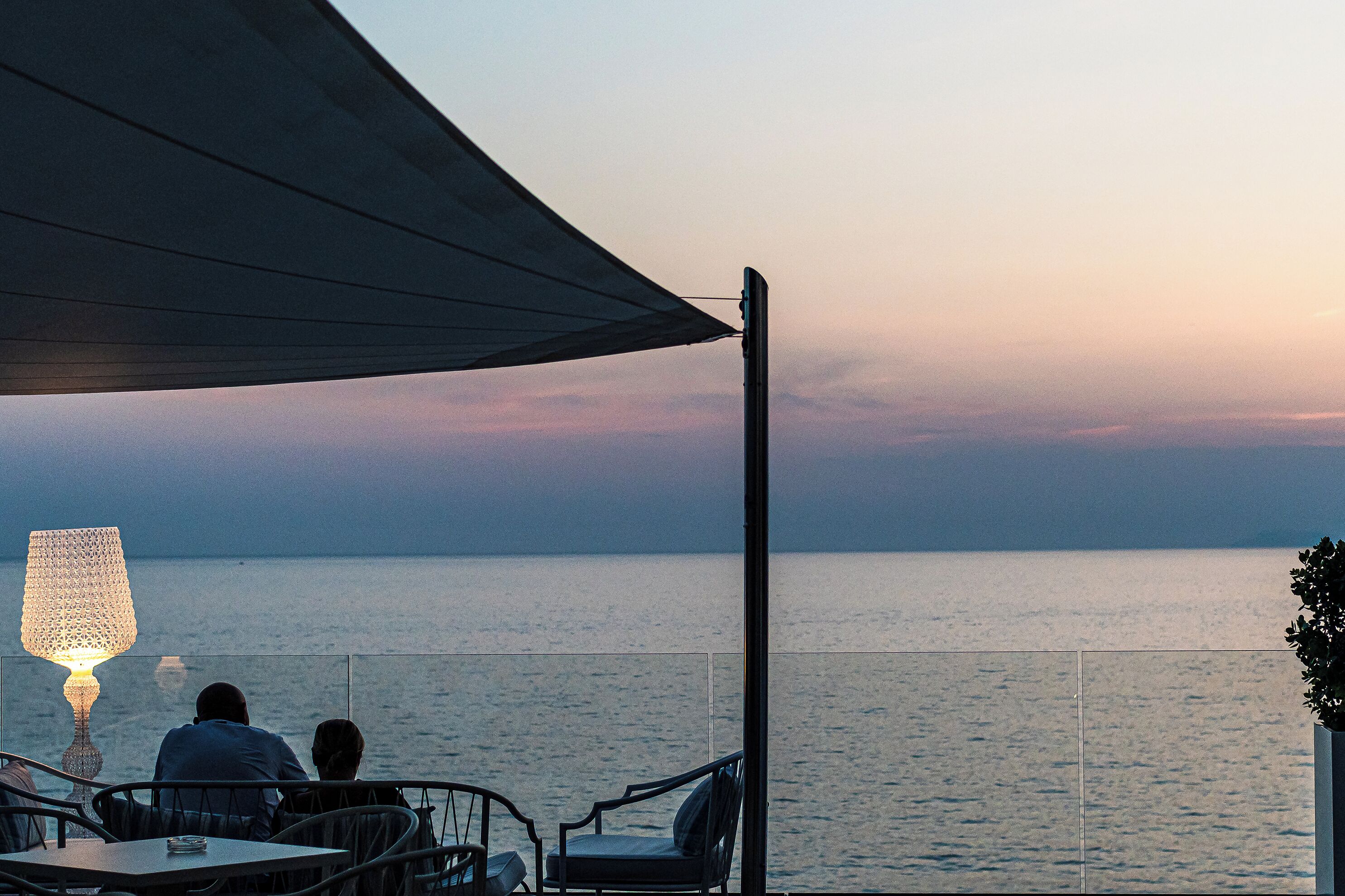 Eine Terrasse mit zwei Stühlen und einem Tisch, mit Blick auf ein ruhiges Meer bei Sonnenuntergang.