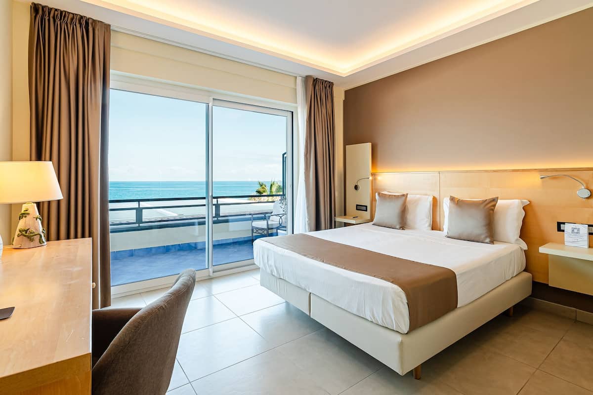 A hotel room with a large bed, a desk, a chair, a nightstand, a lamp, and a view of the ocean through a sliding door.