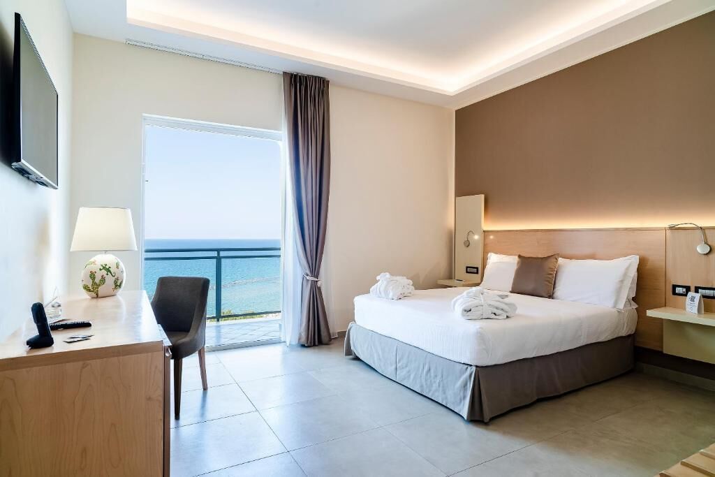 Een hotelkamer met een bed, bureau, stoel en uitzicht op zee.