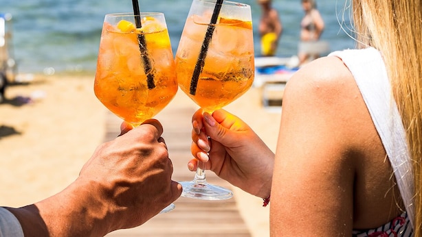 Duas pessoas brindando com bebidas em um deck de madeira à beira-mar.