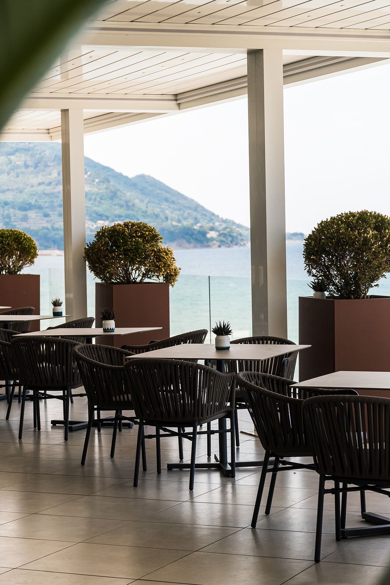 An outdoor seating area with tables and chairs, potted plants, and a view of the sea and mountains.