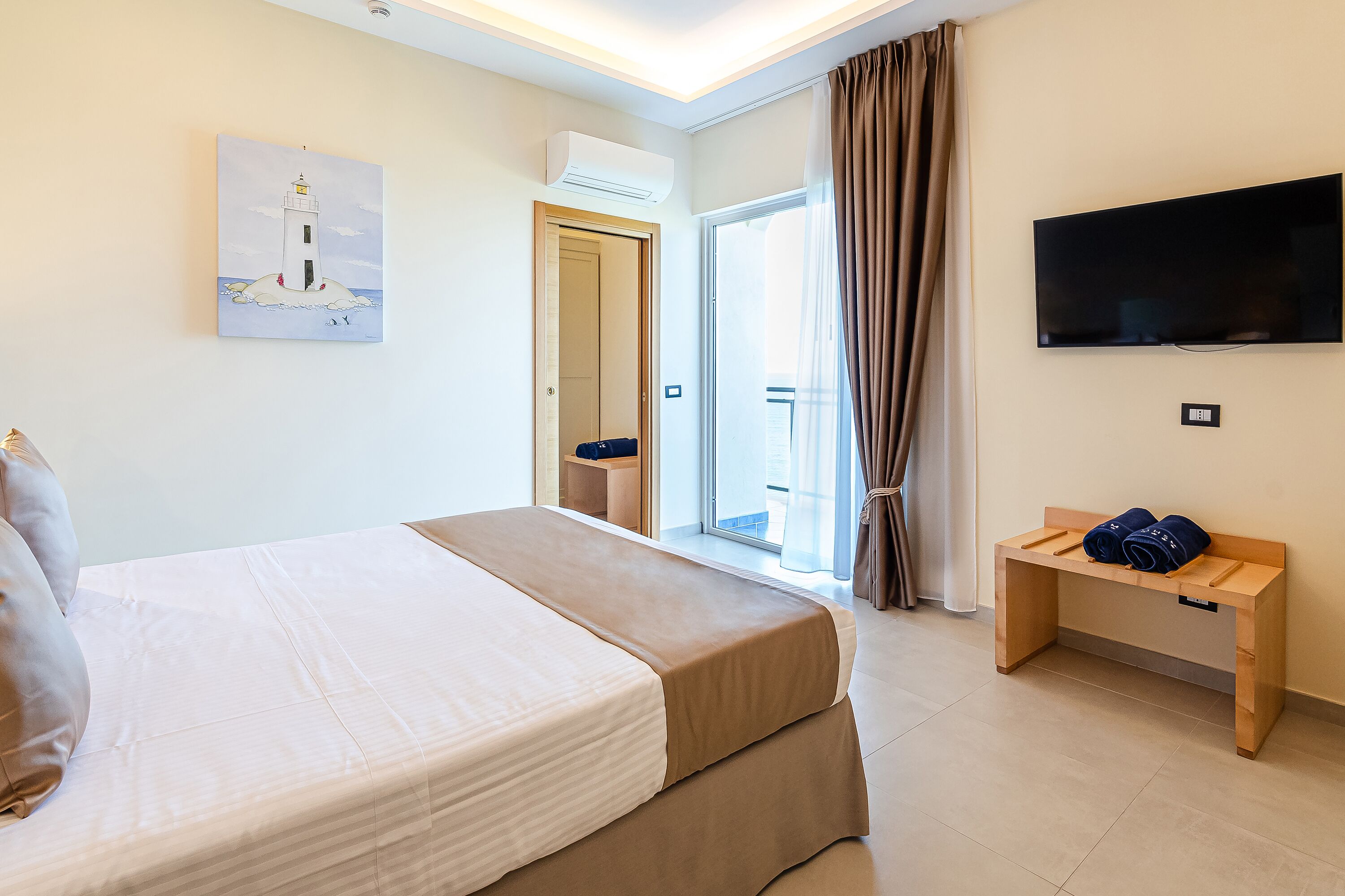 A hotel room with a bed, a TV, a painting, and a bench with towels.