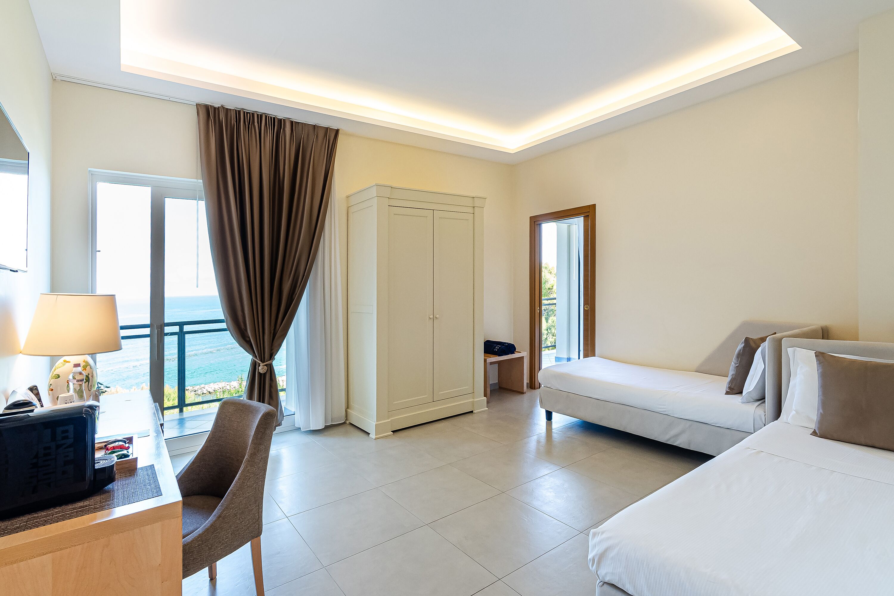 Una camera d'albergo con un letto, una scrivania con una lampada, una sedia, un armadio e vista sul mare.