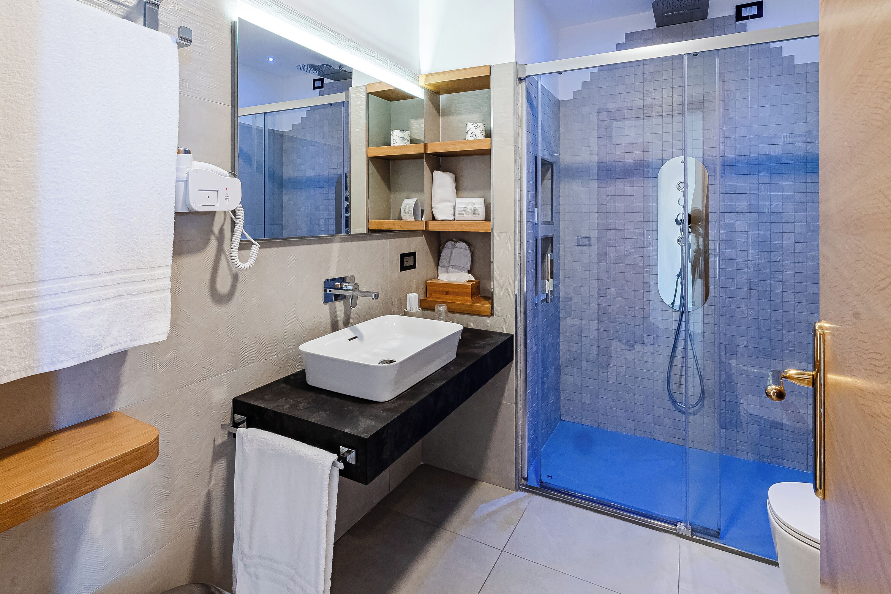 A modern bathroom with a white sink, a glass-enclosed shower, and a wooden shelf with toiletries.