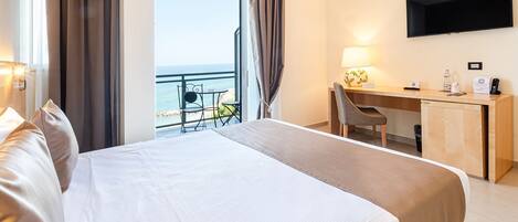 A hotel room with a bed, a desk, a chair, a TV, and a view of the sea.