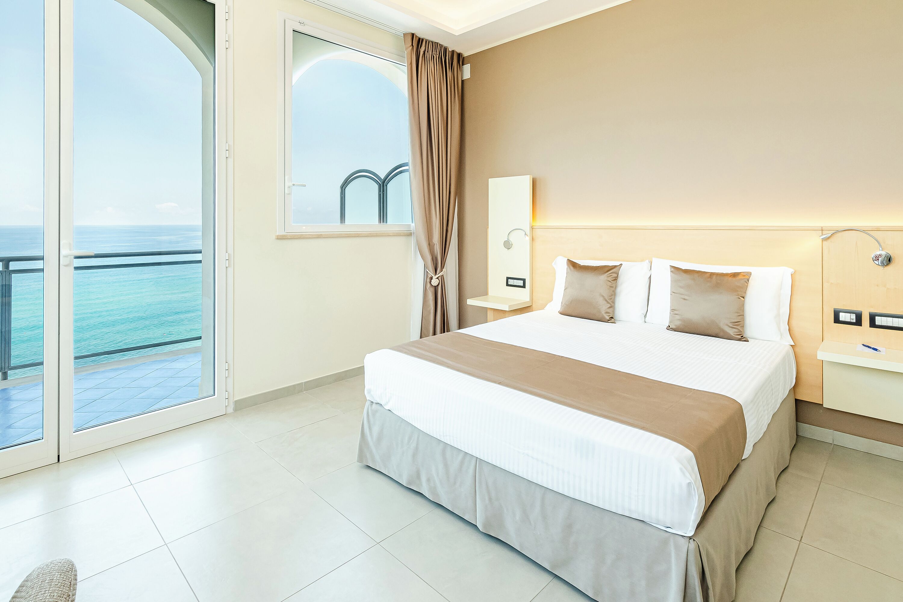 A hotel room with a bed, a nightstand, a mirror, and a view of the ocean through a large window.
