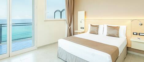 A hotel room with a bed, a nightstand, a mirror, and a view of the ocean through a large window.