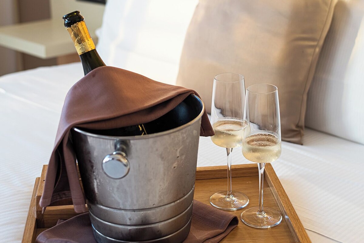 A bed with a bedside table holding a bottle of champagne, two glasses, and a tray.