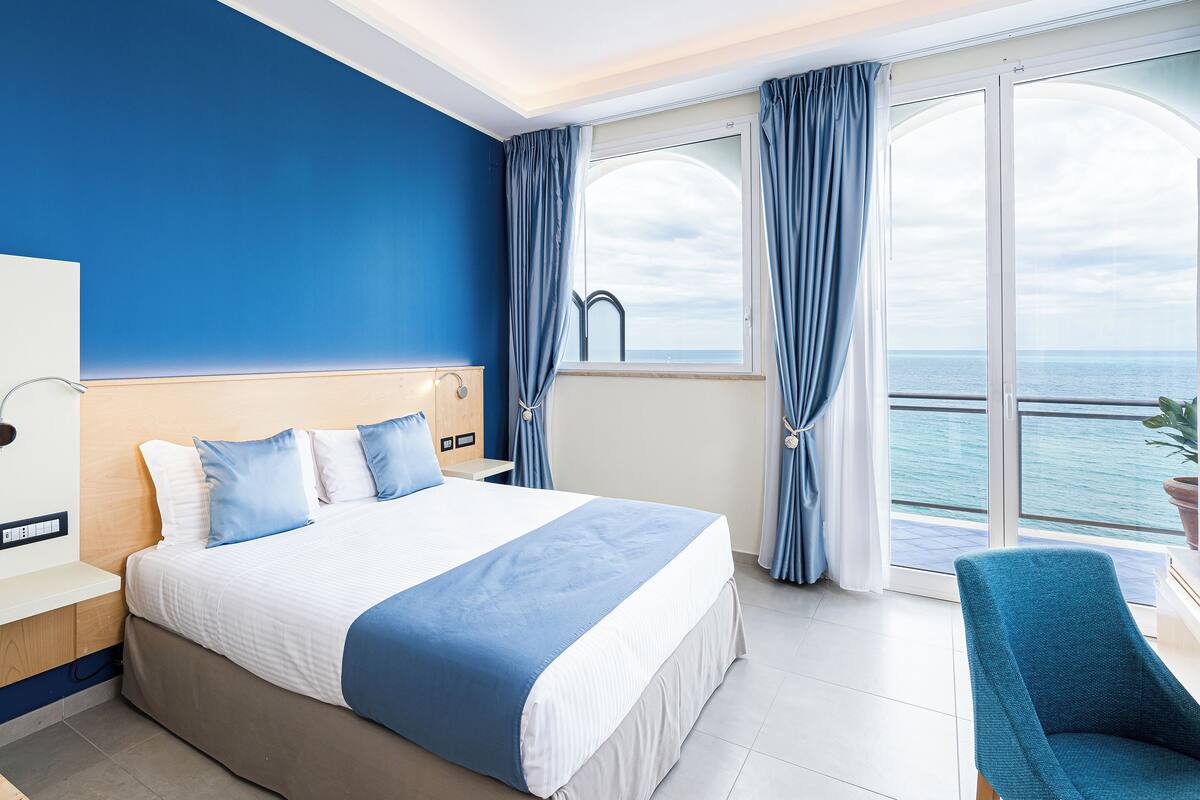 Ein Hotelzimmer mit einem großen Bett, einem Schreibtisch und einem Sessel. Ein Fenster mit Vorhängen bietet Blick auf das Meer.