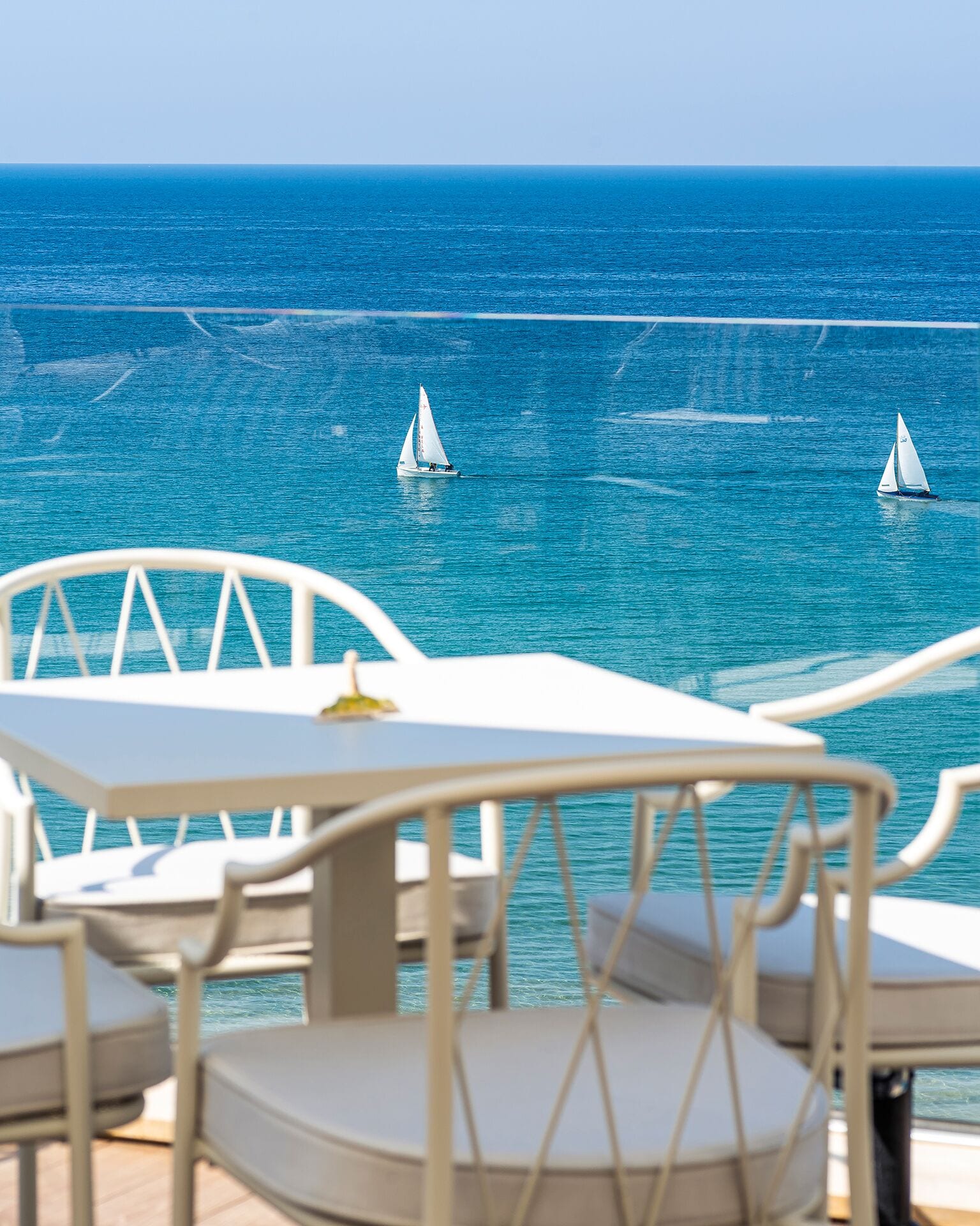A terrace with white furniture overlooking a calm sea with sailboats.