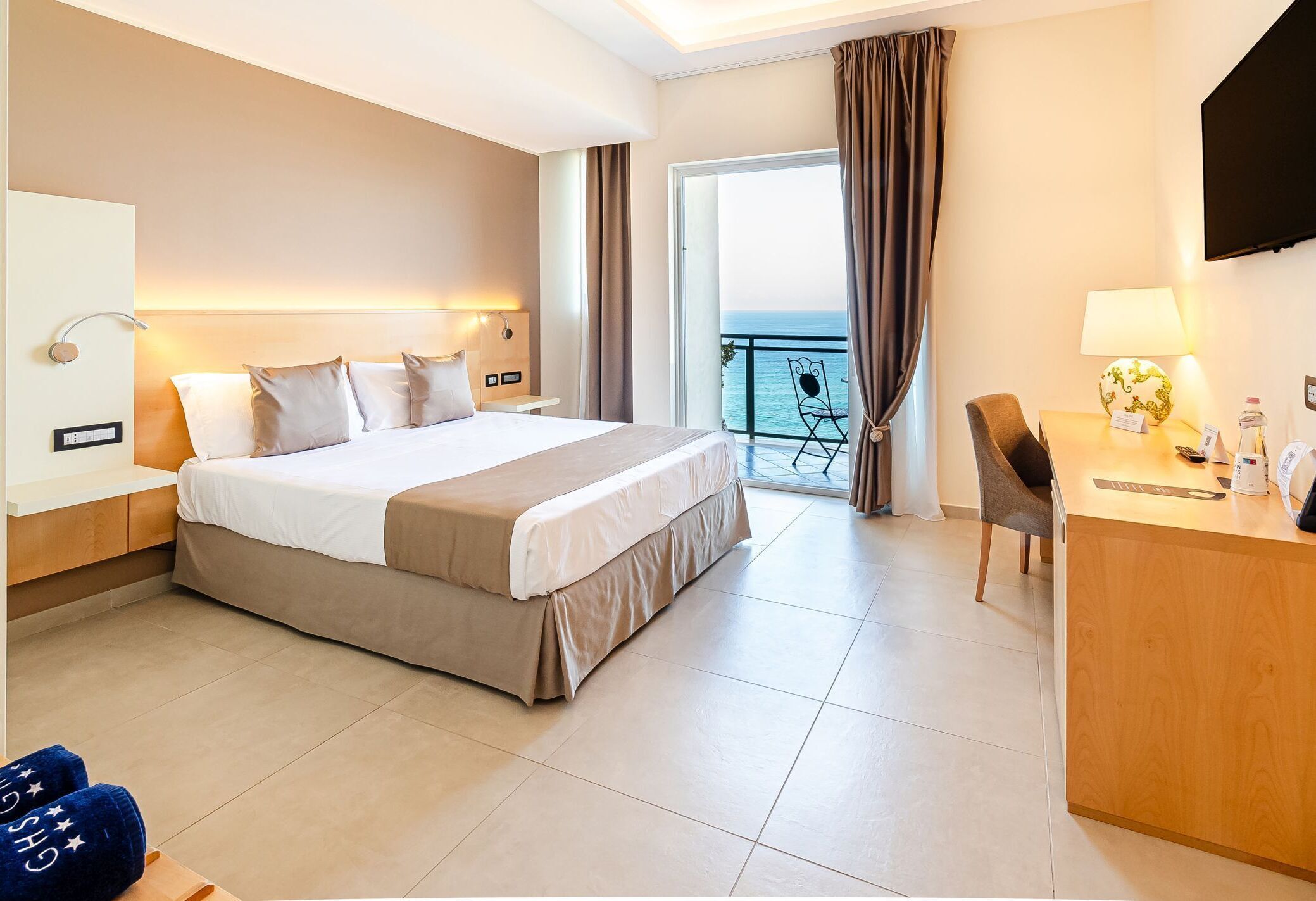 A hotel room with a bed, a desk, a chair, a TV, and a balcony with a view.