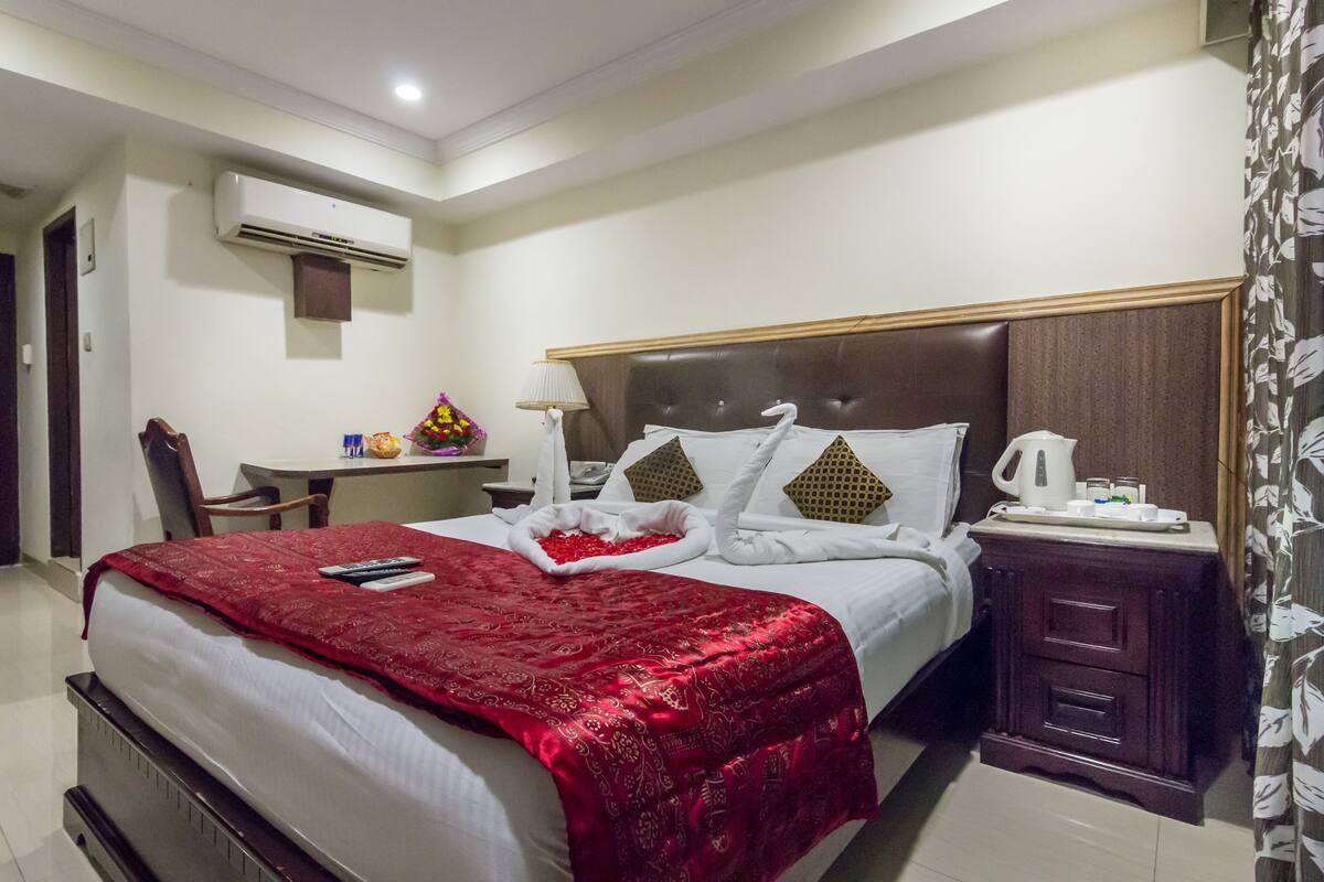 A hotel room with a neatly made bed, a bedside table with a lamp, a small desk with a chair, and an air conditioning unit on the wall.