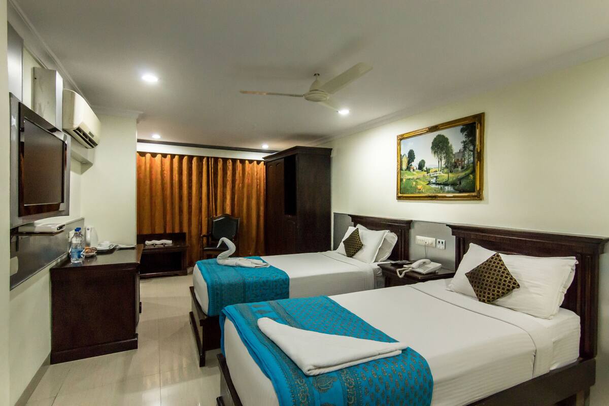 A hotel room with two beds, a desk, a chair, an air conditioning unit, a painting on the wall, and a ceiling fan.