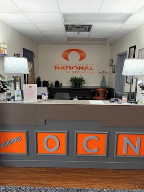 A reception desk at Orange County National Golf Center and Lodge with a 'Welcome' sign, a plant, and a 'germ' sanitizer dispenser. - Orange County National Lodge (Winter Garden)