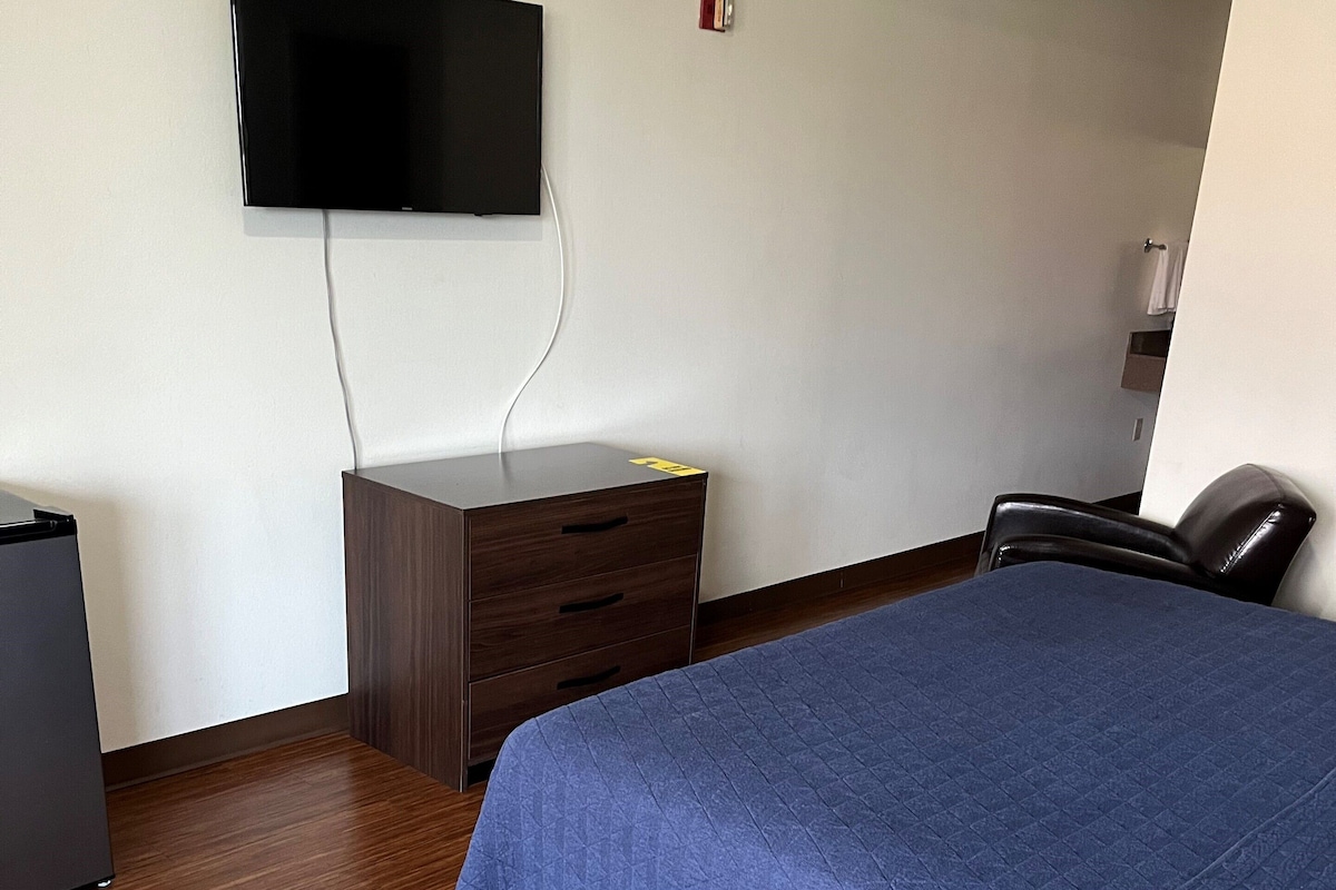 A hotel room with a bed, a desk, a chair, and a flat-screen TV mounted on the wall.