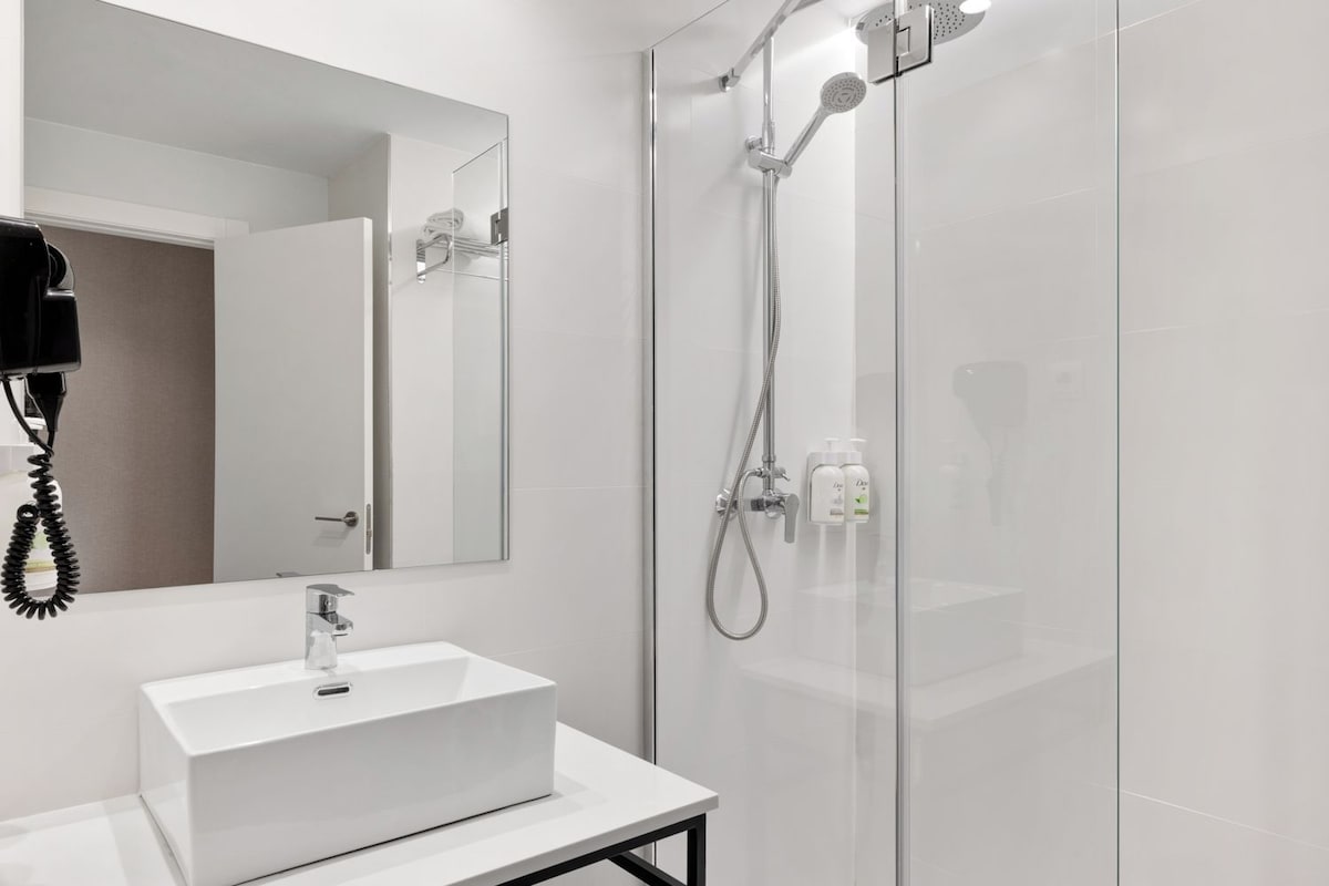 Une salle de bain moderne avec un lavabo blanc, un miroir, une douche en verre et un sèche-cheveux fixé au mur.