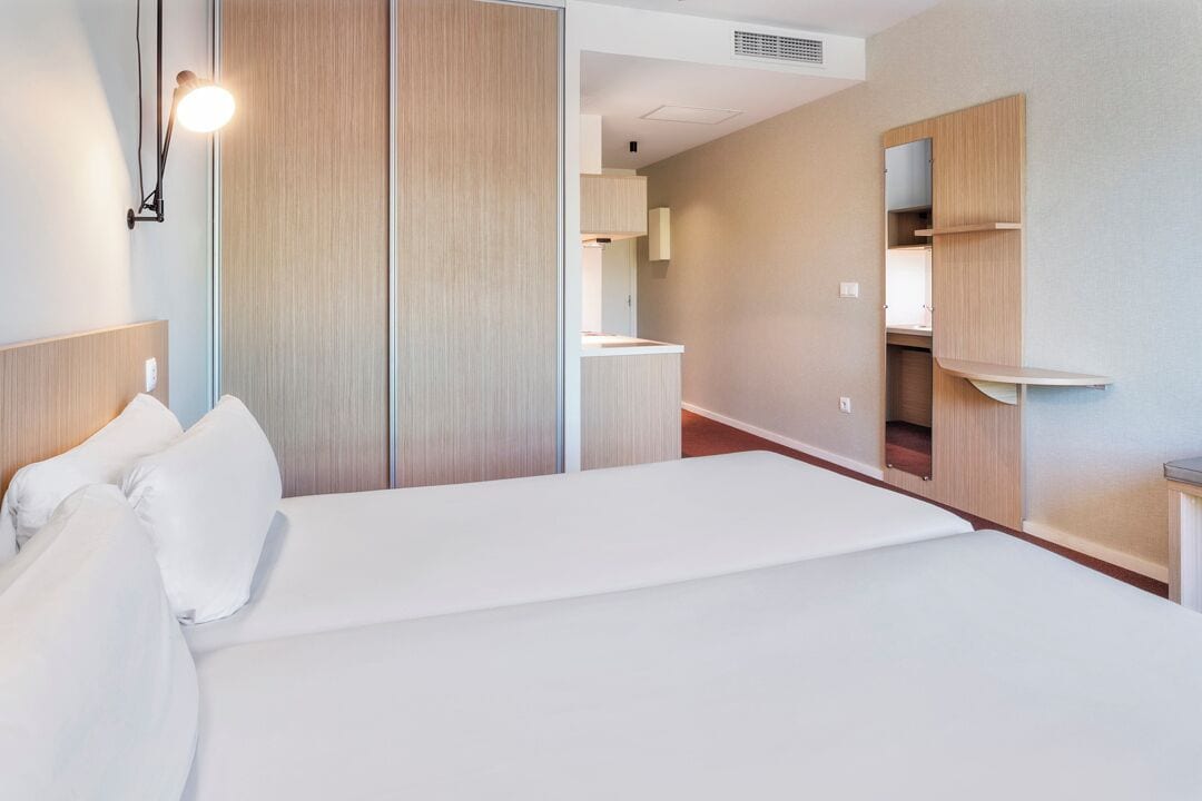Una habitación de hotel con una cama, una mesita de noche, un espejo, un armario y una pequeña zona de kitchenette.