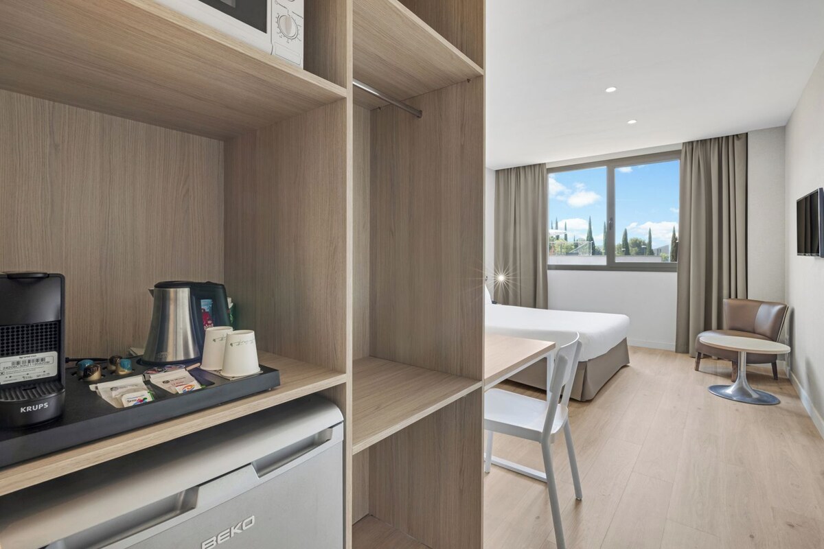 A hotel room with a bed, a desk with a coffee maker, a chair, and a view of the outdoors.