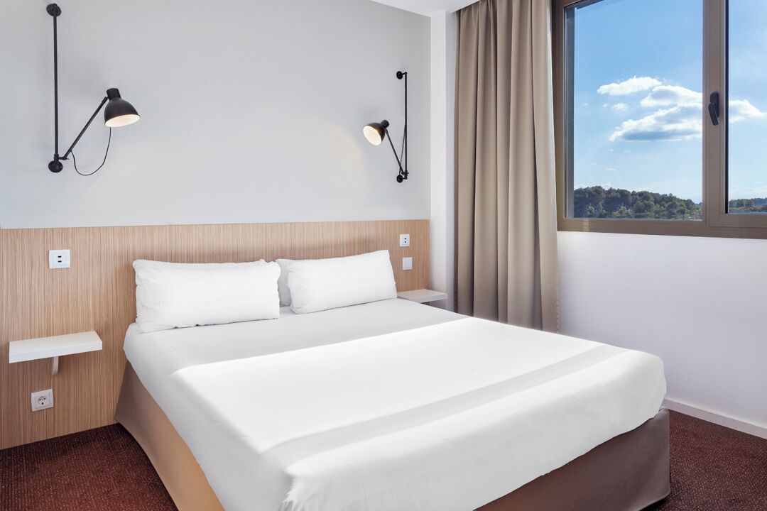 Habitación de hotel con cama, mesitas de noche, lámparas de pared, una ventana con cortinas y vistas al exterior.