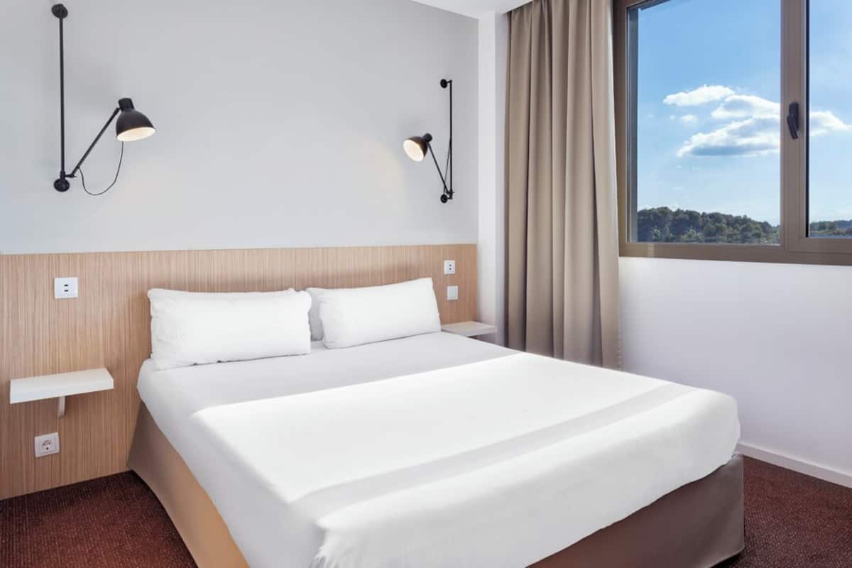 A hotel room with a bed, bedside tables, wall-mounted lamps, a window with curtains, and a view of the outdoors.
