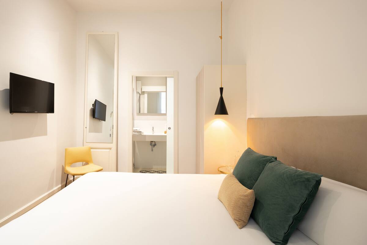 A bedroom with a bed, a chair, a TV, a mirror, and a bathroom visible through an open door.