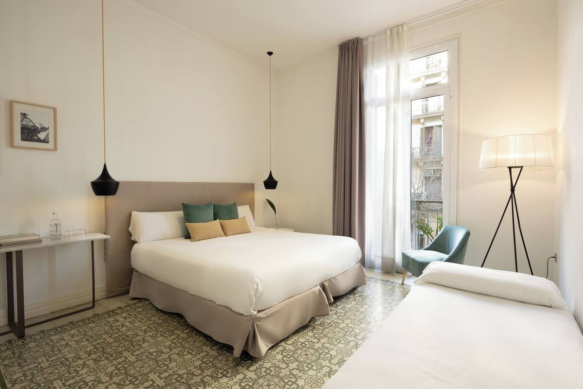 A hotel room with a large bed, a bedside table with a lamp, a chair, and a view of the city through the window.
